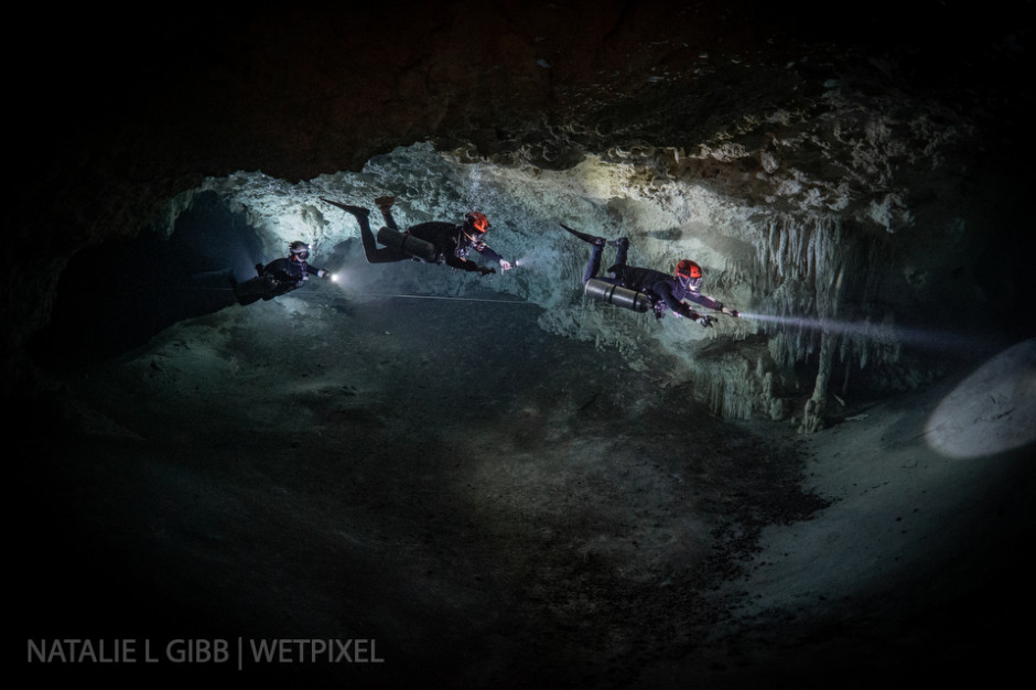 Three divers float in a line, with video lights placed in front and behind the divers to show the cave.  The models are also carrying their own lights to illuminate their bodies. 